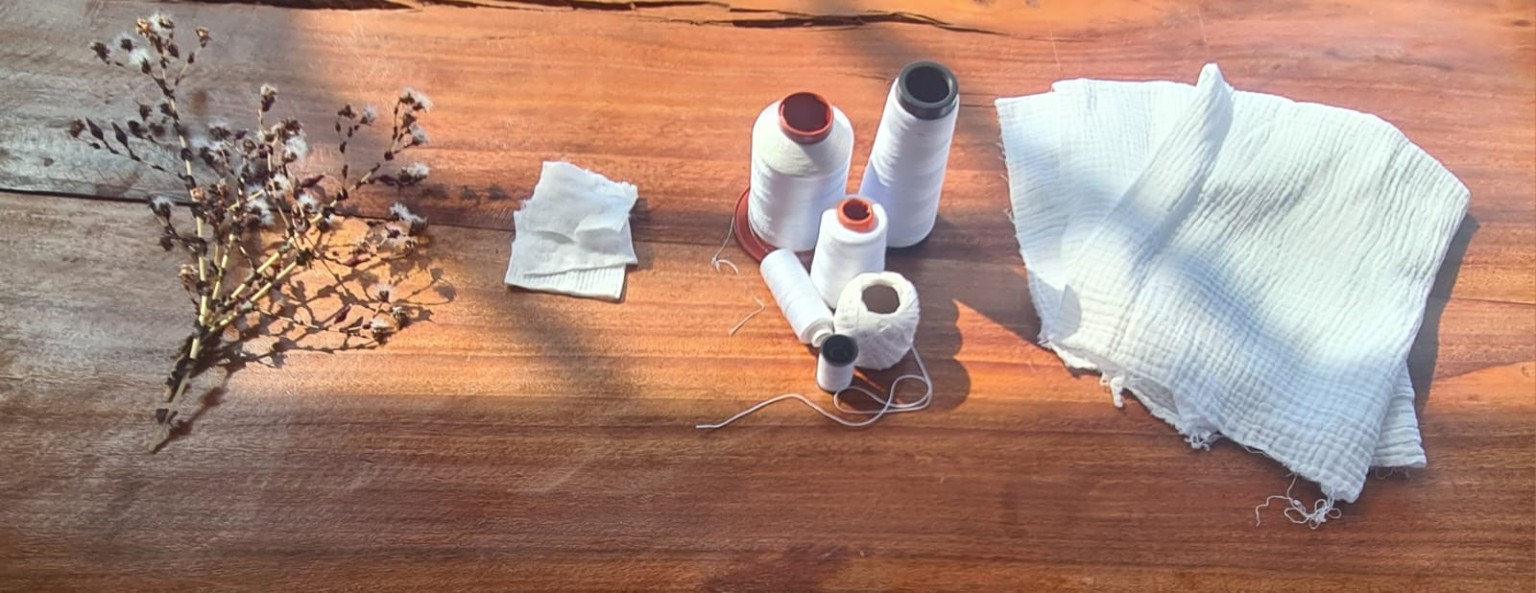 Composição sobre uma mesa de madeira com cones de fio, pequenos recortes de tecido e gaze branca em luz natural.