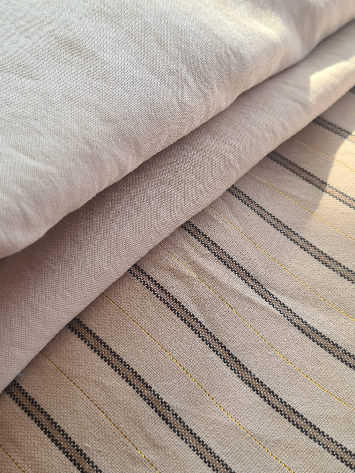 Tecidos neutros dobrados lado a lado, incluindo um tecido listrado com fio dourado e tecidos de superfície lisa.